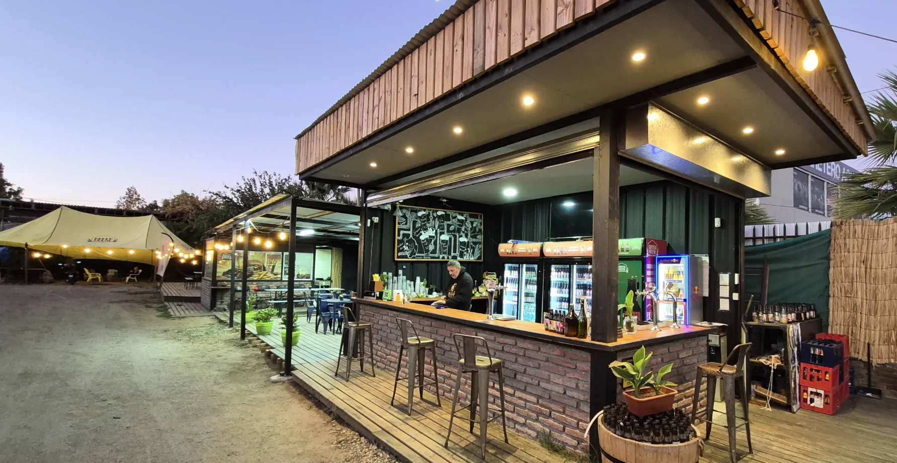 Barra de ladrillo y madera con tiradores de cerveza y refrigeradores, y una terraza de madera con mesas y un toldo con luces, al atardecer