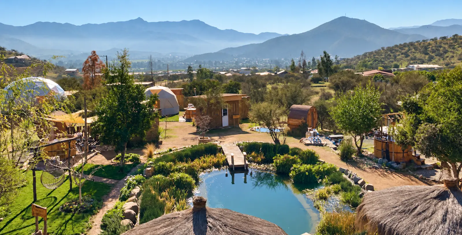 Vista desde lo alto de EcoRiam: domos, cabañas y tinajas de madera, una laguna natural con un muelle y techos de paja, con el valle de Curacaví y la cordillera al fondo